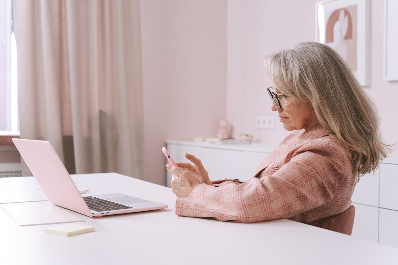 Elderly woman in a pink jacket using a smartphone at a desk with a laptop in a stylish office.
